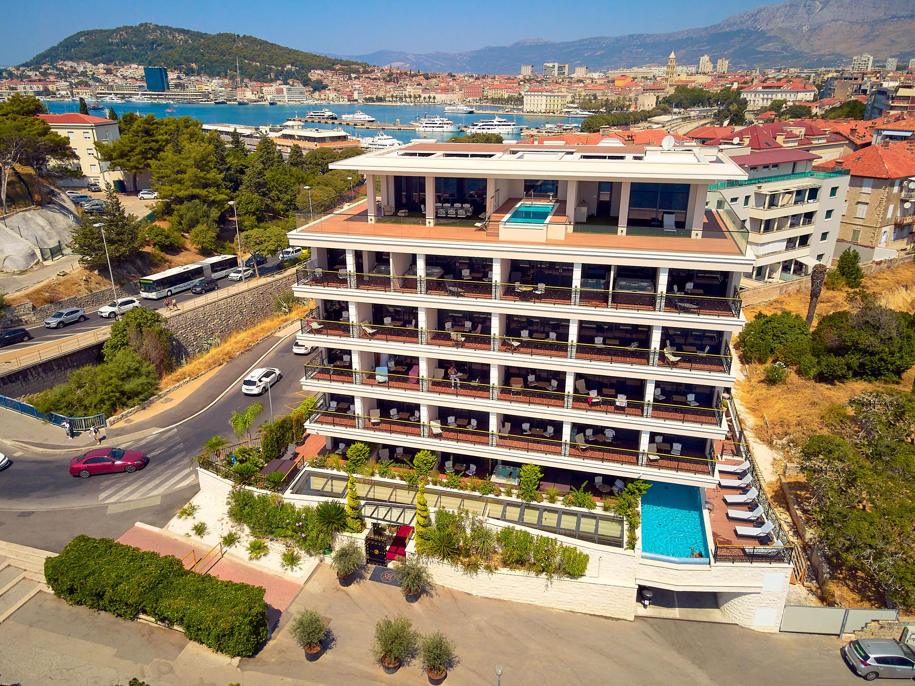 Un hotel de varios pisos con piscina en la azotea, situado cerca de la costa, con embarcaciones a la vista y un paisaje urbano al fondo.