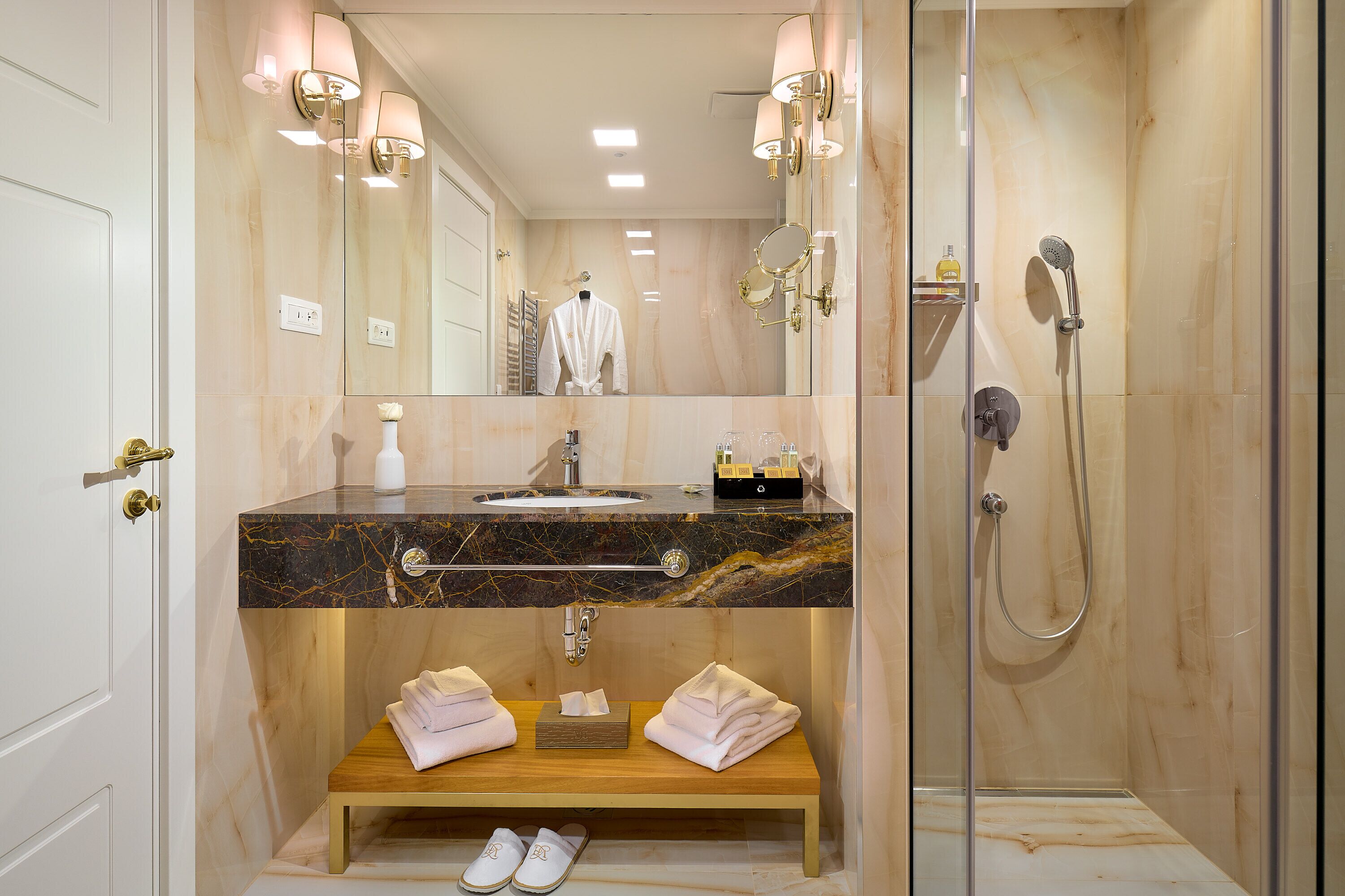 A modern bathroom with a marble vanity, a glass-enclosed shower, and a wooden bench with towels.