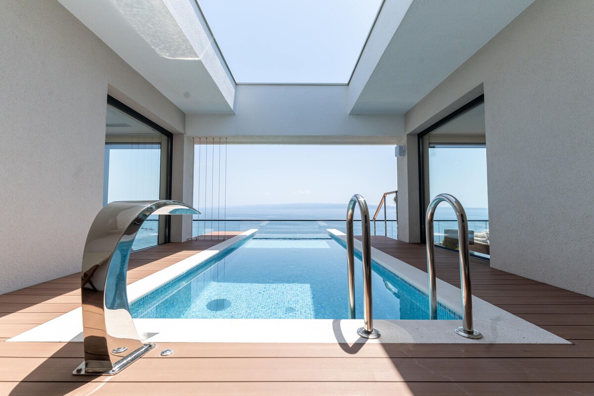 A modern infinity pool with a clear view of the ocean, a skylight above, and a wooden deck.