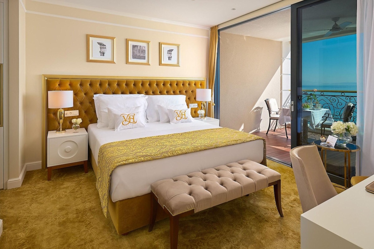 A hotel room with a large bed, a bench, a desk, and a balcony with a view.