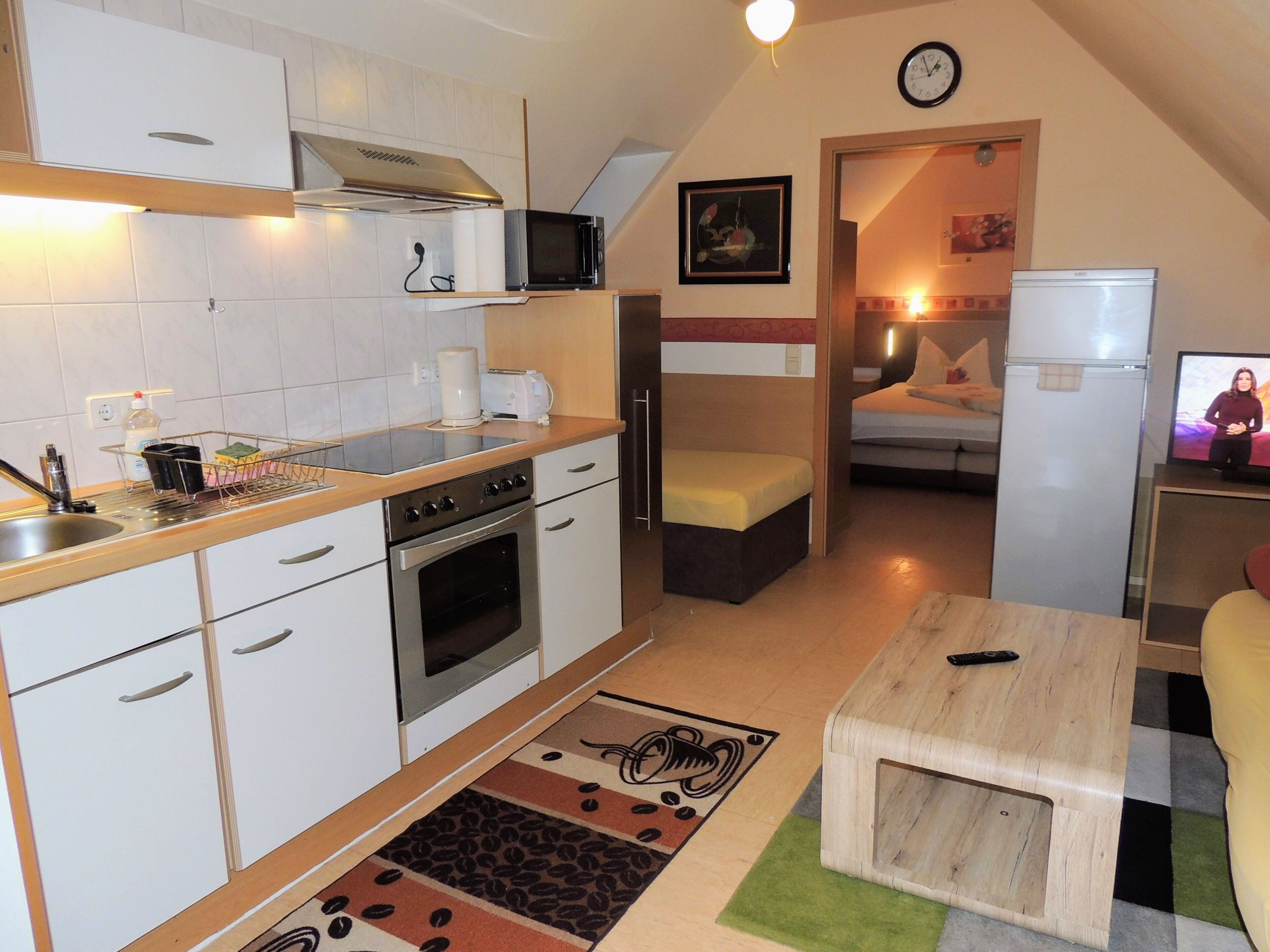 Cocina compacta con microondas, tostadora y horno, un pequeño comedor con mesa y sillas, y un dormitorio con cama y televisión.