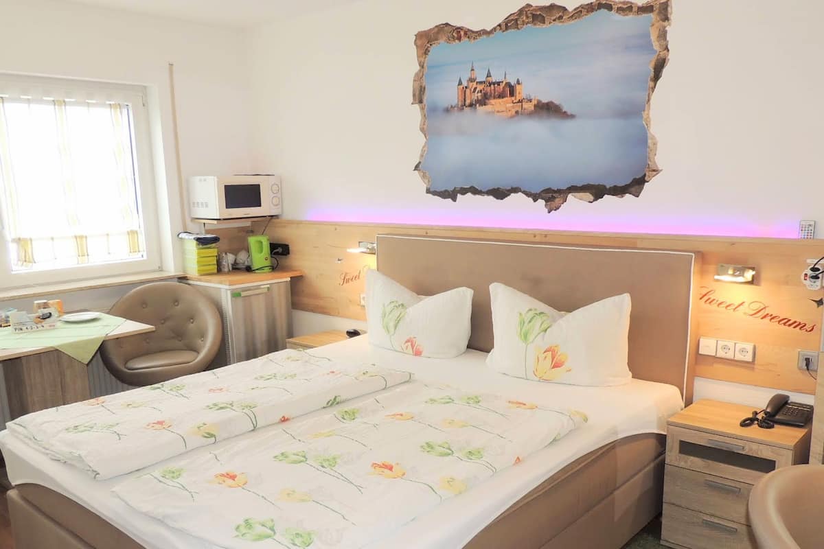A hotel room with a bed, a desk, a chair, a microwave, a kettle, and a painting of a castle.