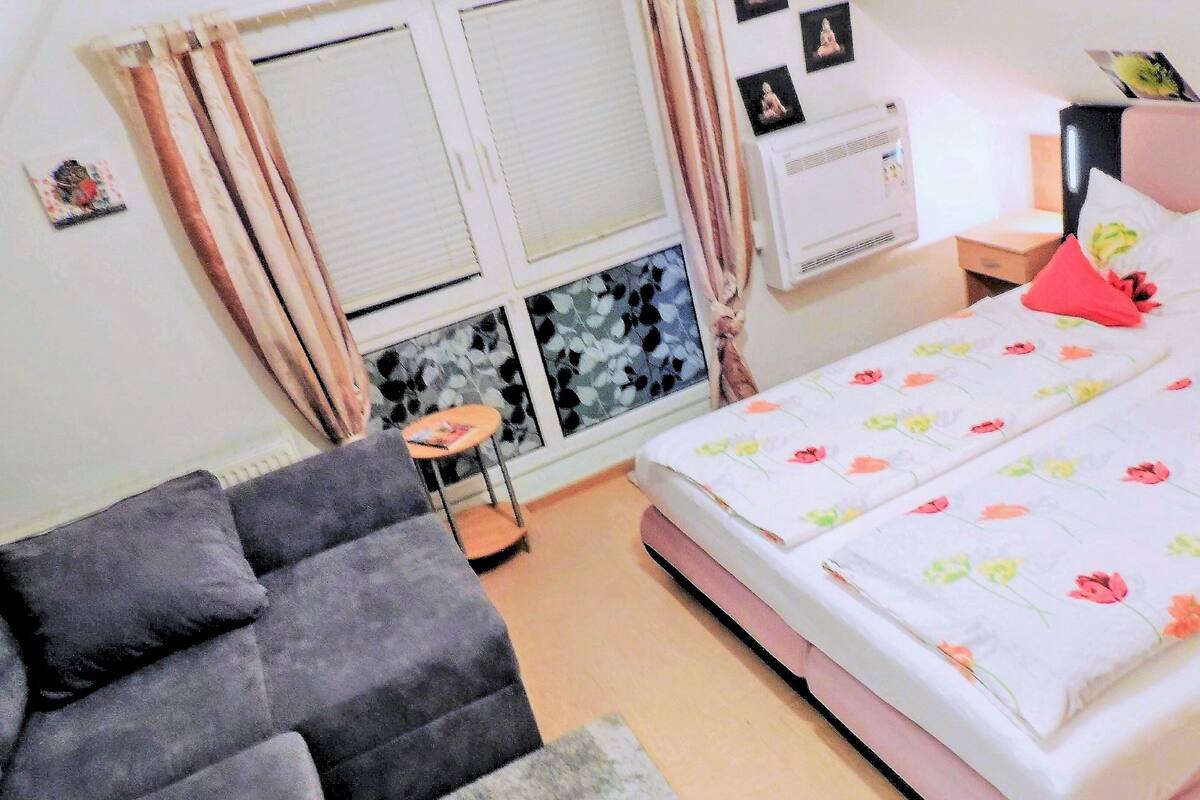 A bedroom with a bed, a small table, a sofa, and a window with curtains.