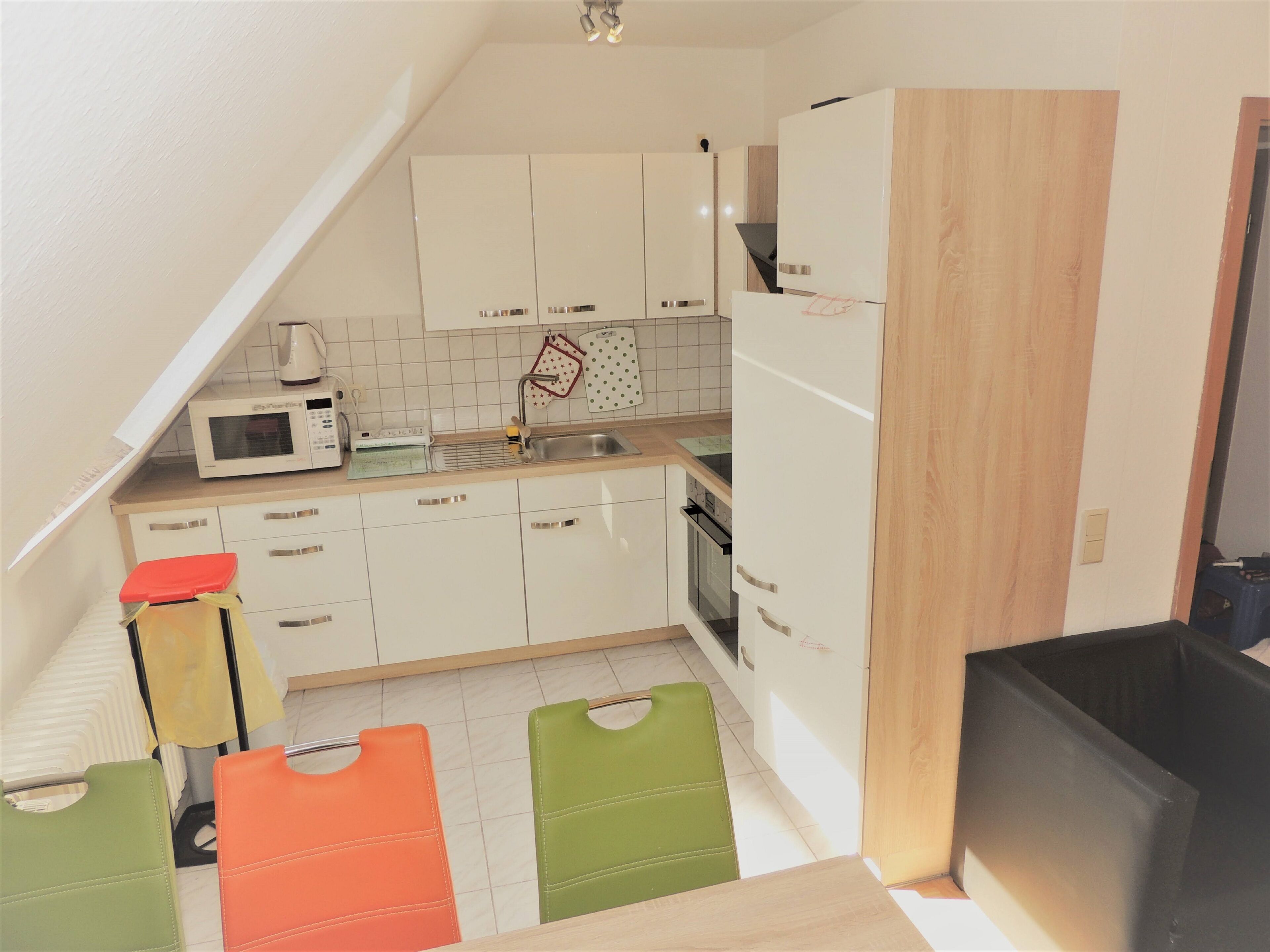 A compact kitchen with white cabinets, a microwave, a sink, and a stove. There are colorful chairs and a black trash bin.