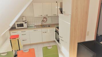A compact kitchen with white cabinets, a microwave, a sink, and a stove. There are colorful chairs and a black trash bin.