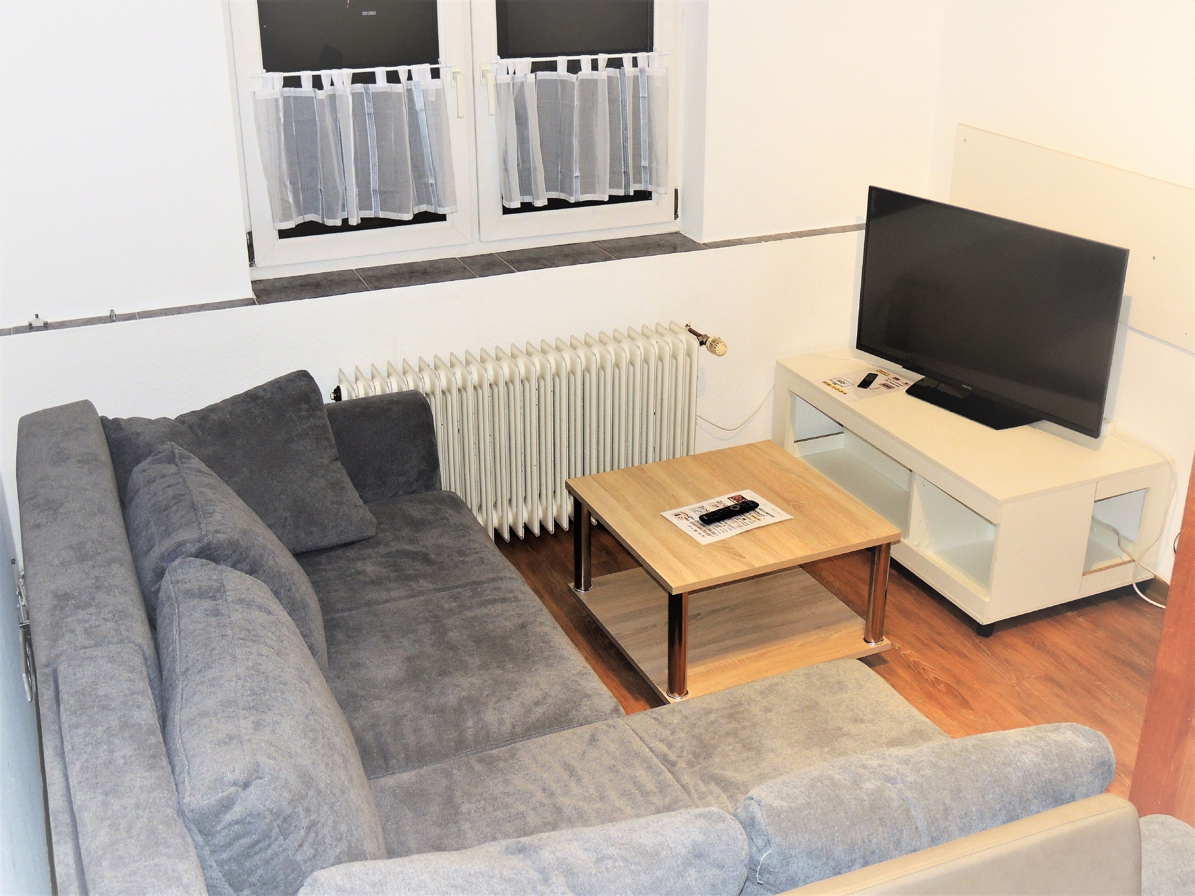 A living room with a grey sofa, a wooden coffee table, a TV on a white stand, and a radiator.