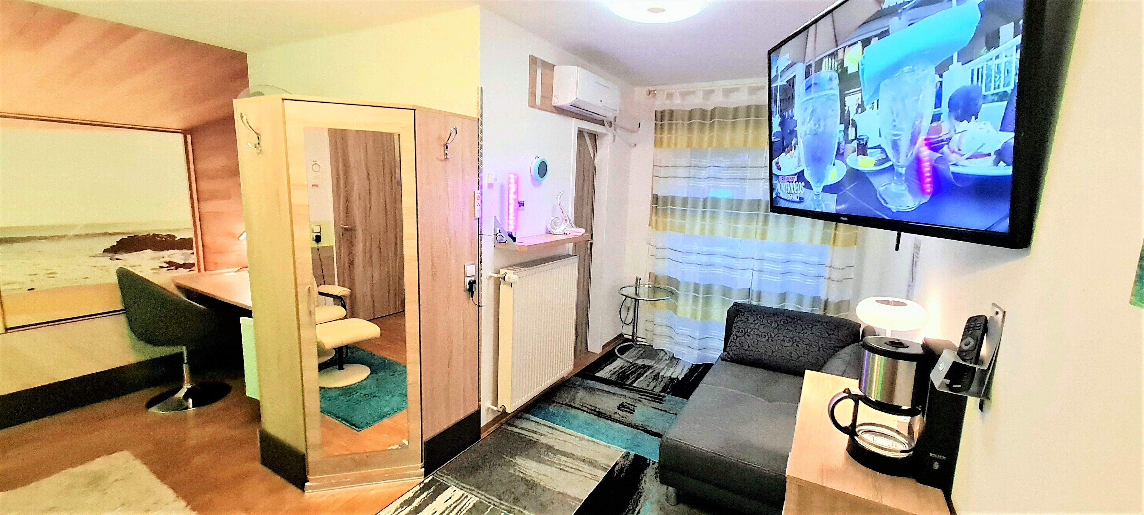 A compact hotel room with a wooden door, a small bathroom, a sofa, a coffee machine, and a TV mounted on the wall.