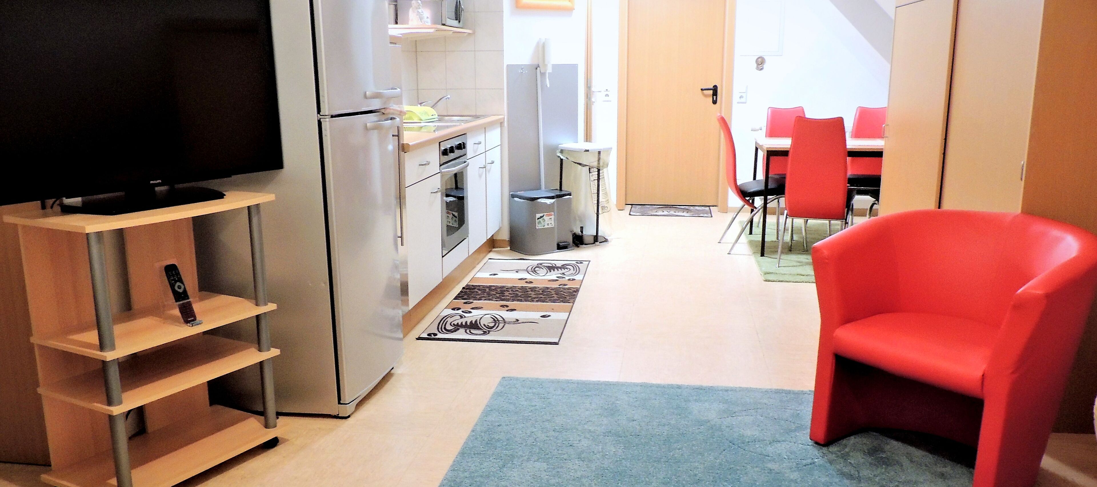 A compact kitchen with a refrigerator, microwave, and oven. There is a dining area with red chairs and a table. A TV is mounted on the wall.