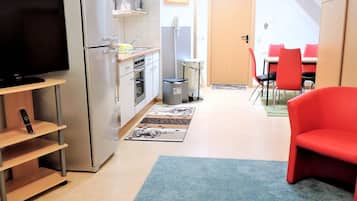 Cocina compacta con refrigerador, microondas y horno. Hay una zona de comedor con sillas rojas y una mesa. Un televisor está montado en la pared.