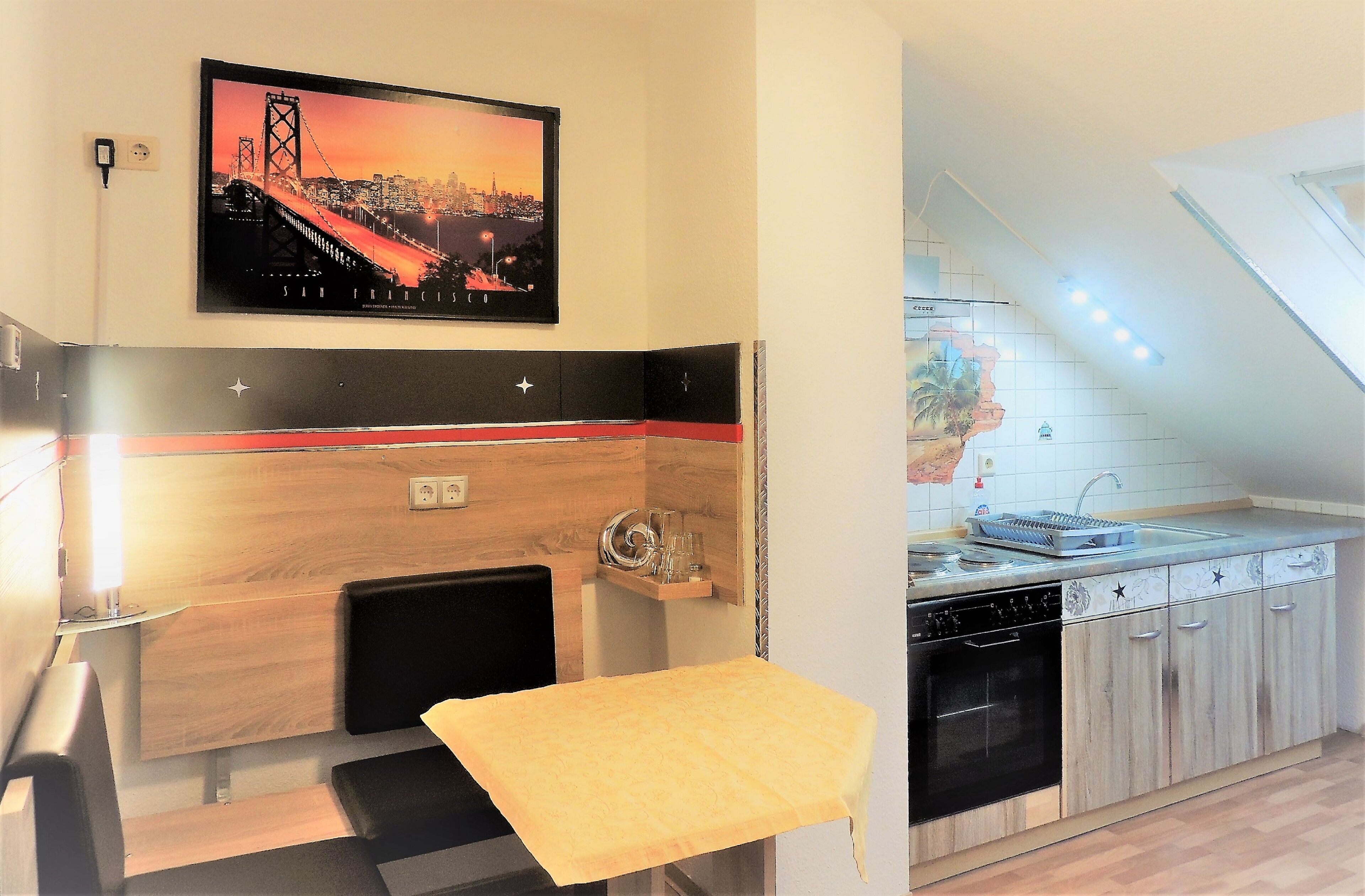 Una cocina compacta con encimera de madera, un lavabo y lavavajillas. Hay una mesa pequeña con mantel amarillo y una foto enmarcada de un puente en la pared.