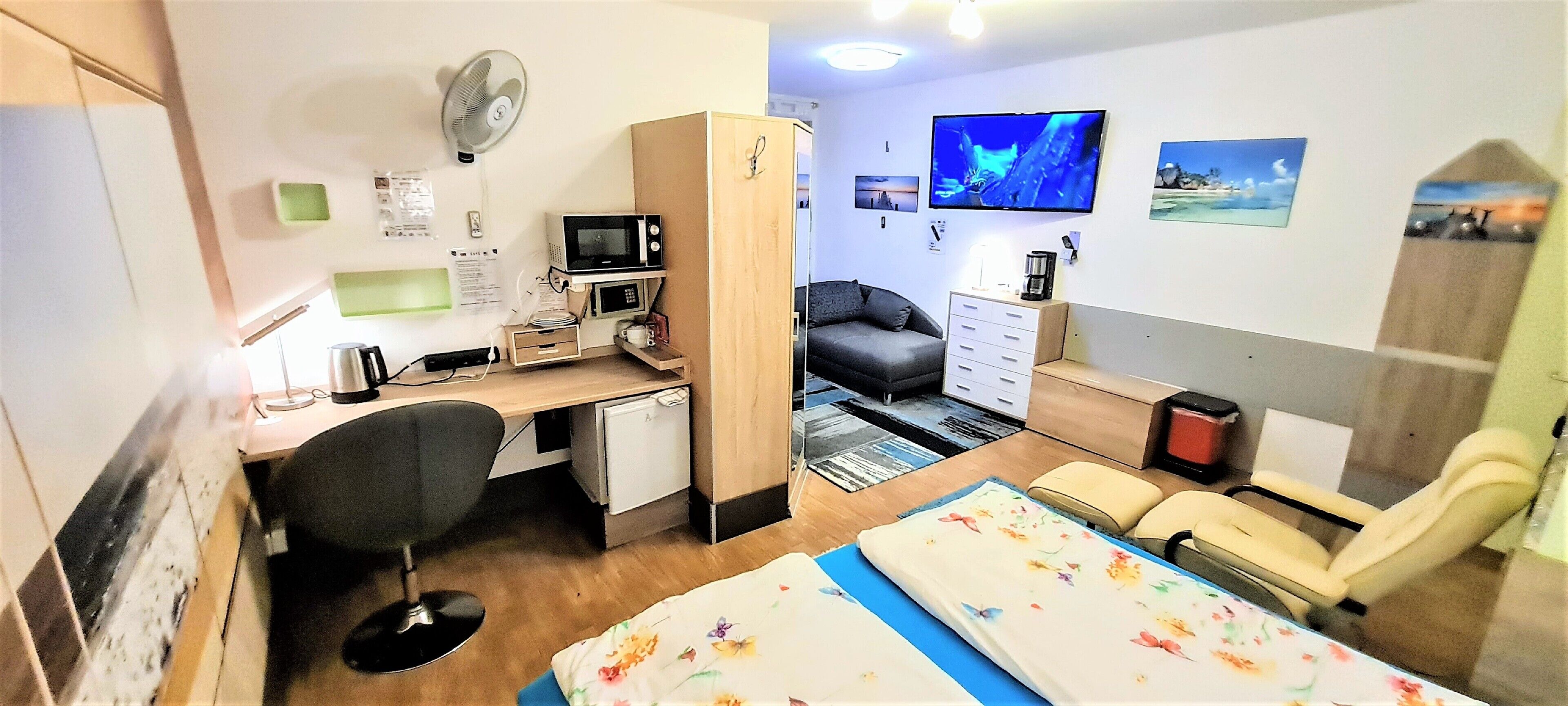 Ein gemütlicher Raum mit Schreibtisch, Stuhl, Bett, Fernseher und einer kleinen Küchenzeile.