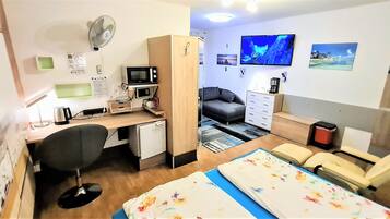 Ein gemütlicher Raum mit Schreibtisch, Stuhl, Bett, Fernseher und einer kleinen Küchenzeile.