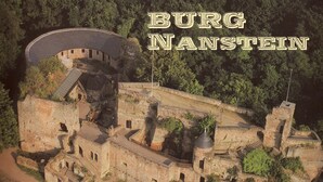 Eine Luftaufnahme von Burg Nanstein, die ihre historische Steinarchitektur, einen Rundturm und einen Innenhof mit Außenbestuhlung zeigt.