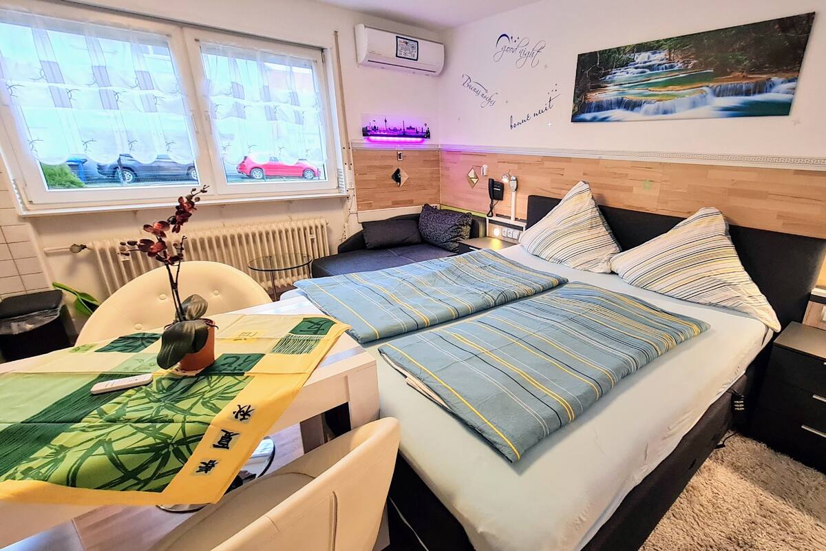 A bedroom with a bed, bedside tables, a desk, and a chair. There is a window with curtains, a radiator, and a wall-mounted air conditioner.