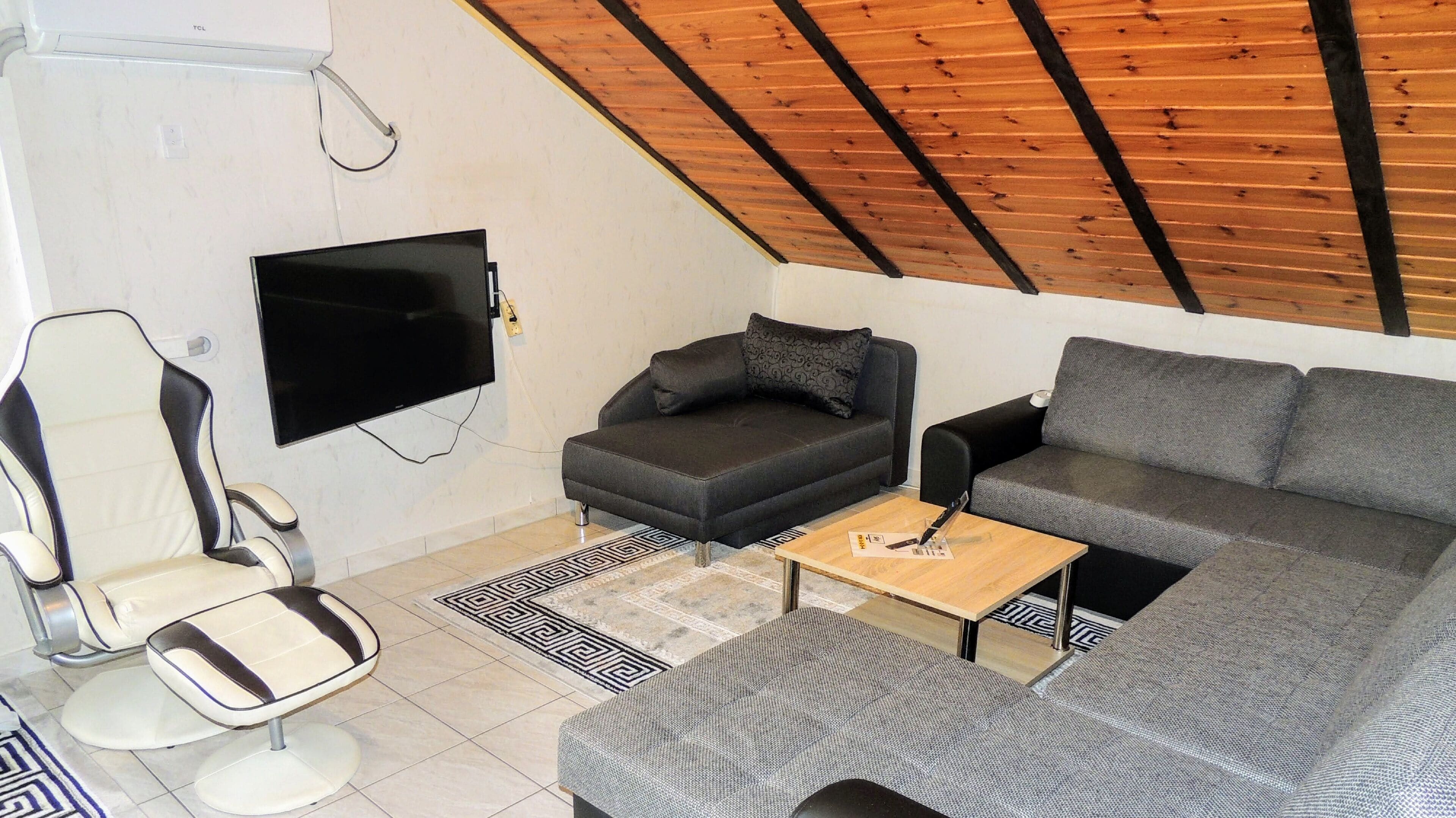 A modern living room with a flat-screen TV mounted on the wall, a grey sofa, a black and white armchair, and a small wooden coffee table.