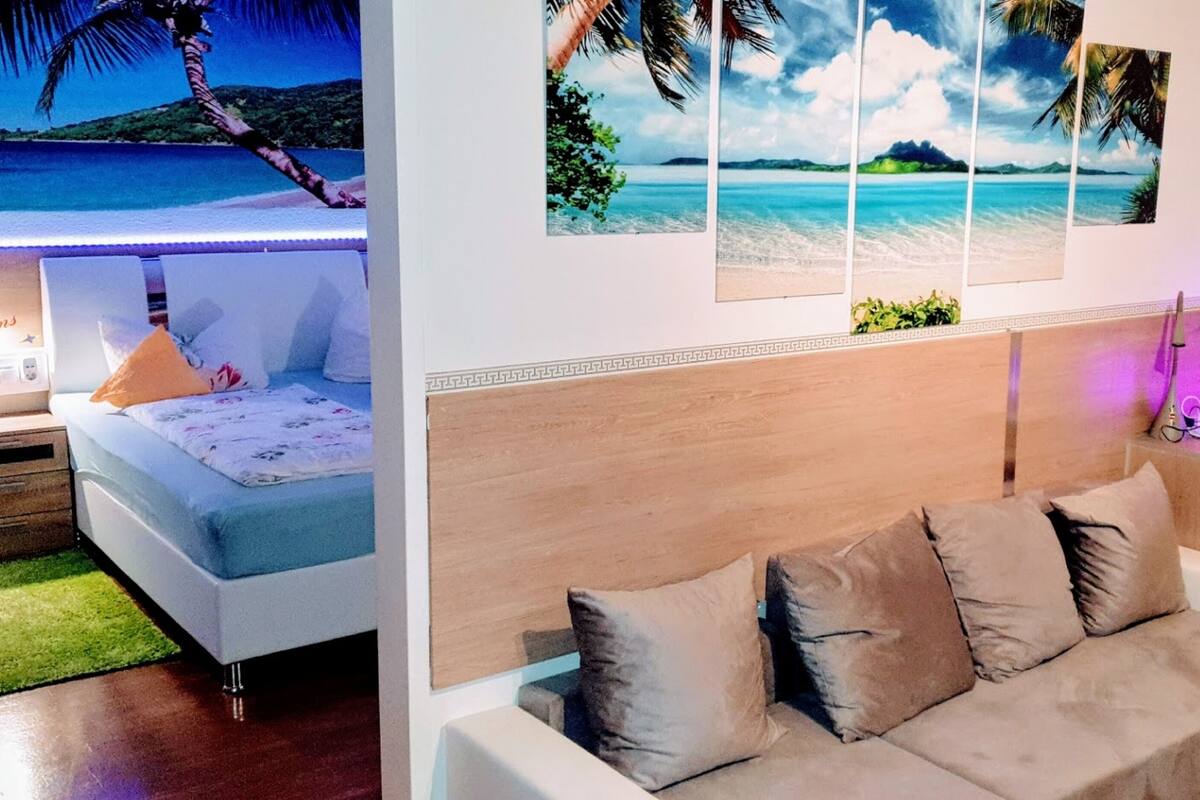 A bedroom with a bed, a green chair, a sofa, and a wall with a beach mural.