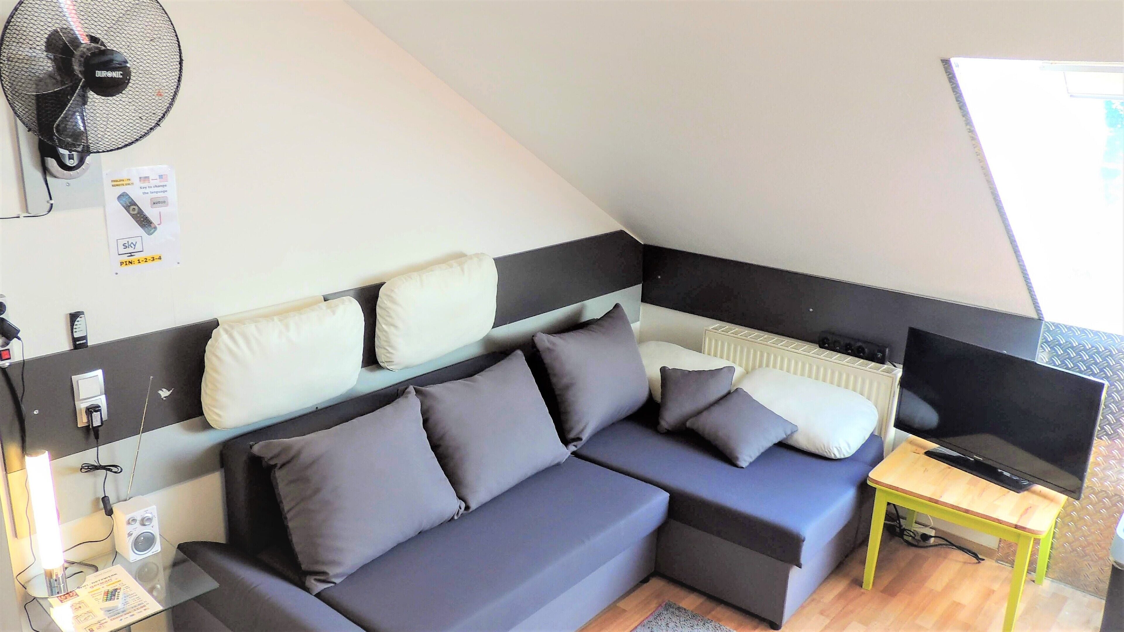 Een moderne, compacte kamer met een modulaire bank, een klein tafeltje, een televisie en een ventilator aan de muur.