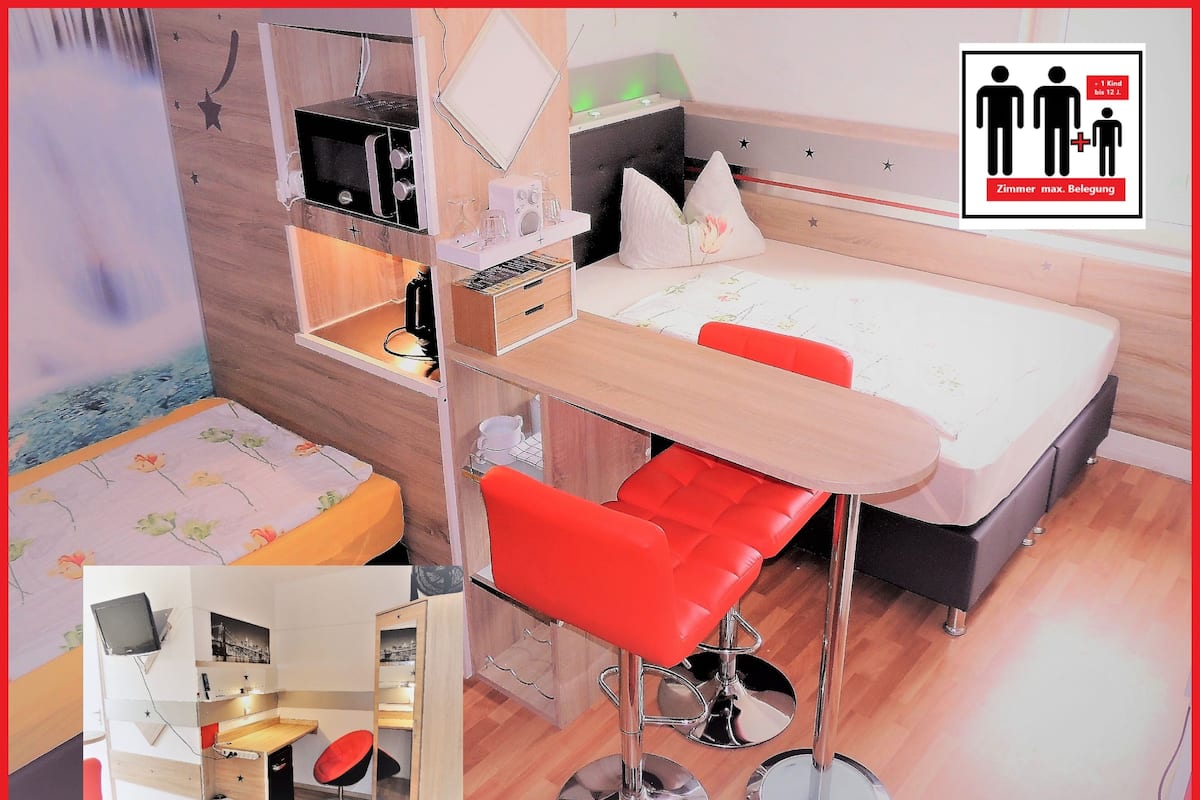 A compact room with a bed, a desk, and a chair. There is a small shelf with a radio and glasses.
