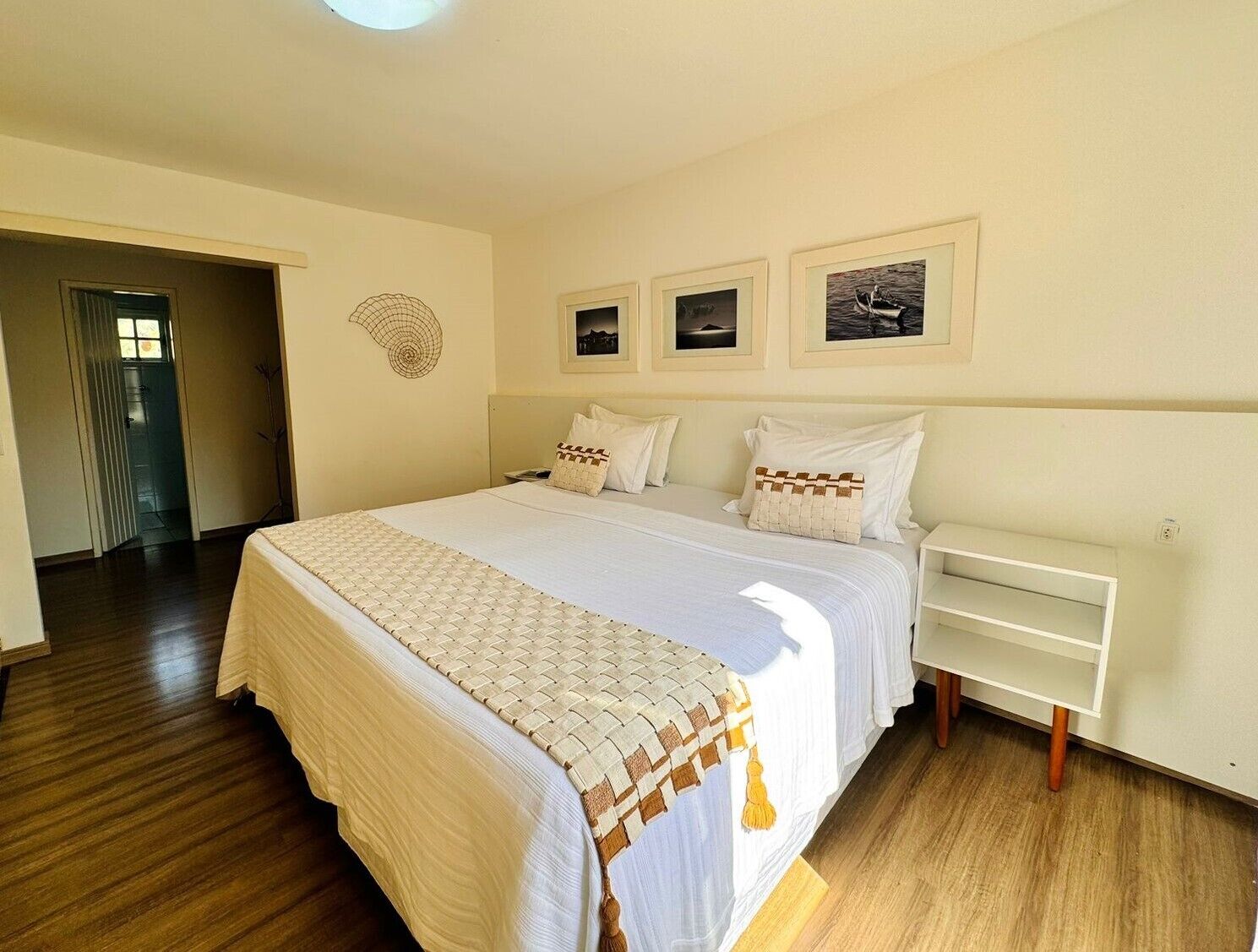 Un dormitorio con una cama grande, una mesita de noche y un estante fijado a la pared. Se ve una puerta que da a otra habitación y una decoración en la pared.