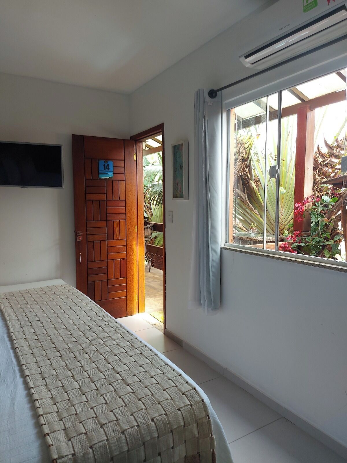 A room with a bed, a wooden door, a window with a view of plants, and an air conditioning unit.