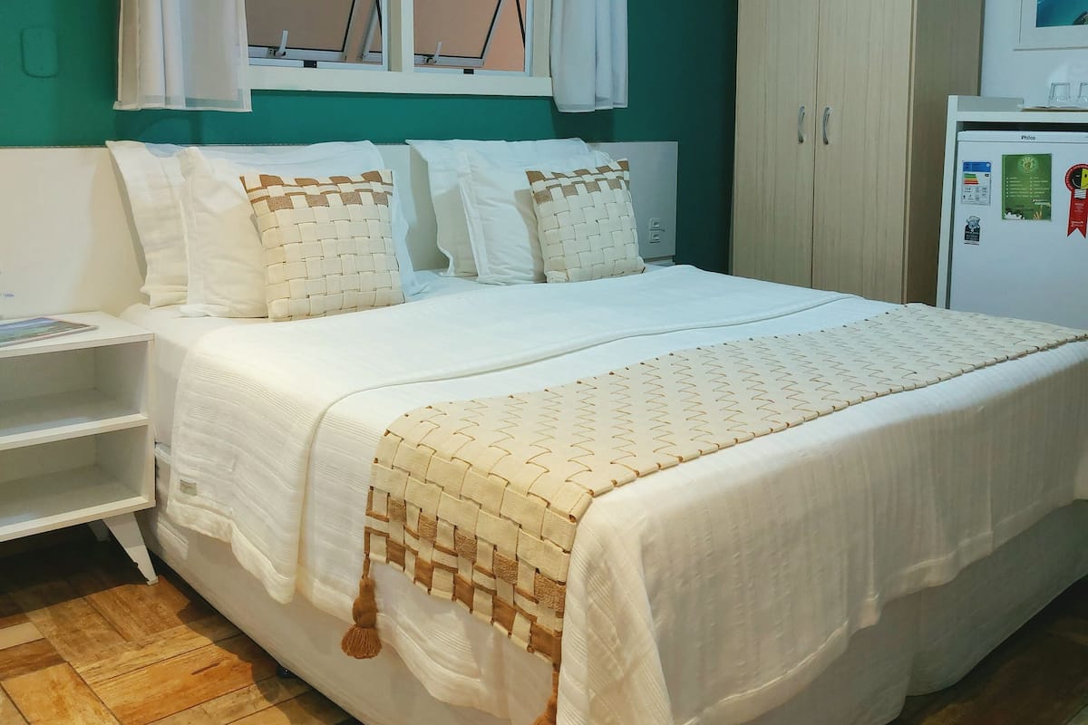 A bedroom with a bed, a window with curtains, a wardrobe, and a refrigerator.