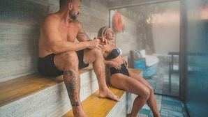 Una pareja sentada en un banco de madera en una habitación con pared de vidrio, un chaleco salvavidas a la vista y una silla azul.