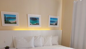 Una cama bien hecha con edredón blanco, una mesita de noche con una planta pequeña y tres cuadros en la pared.