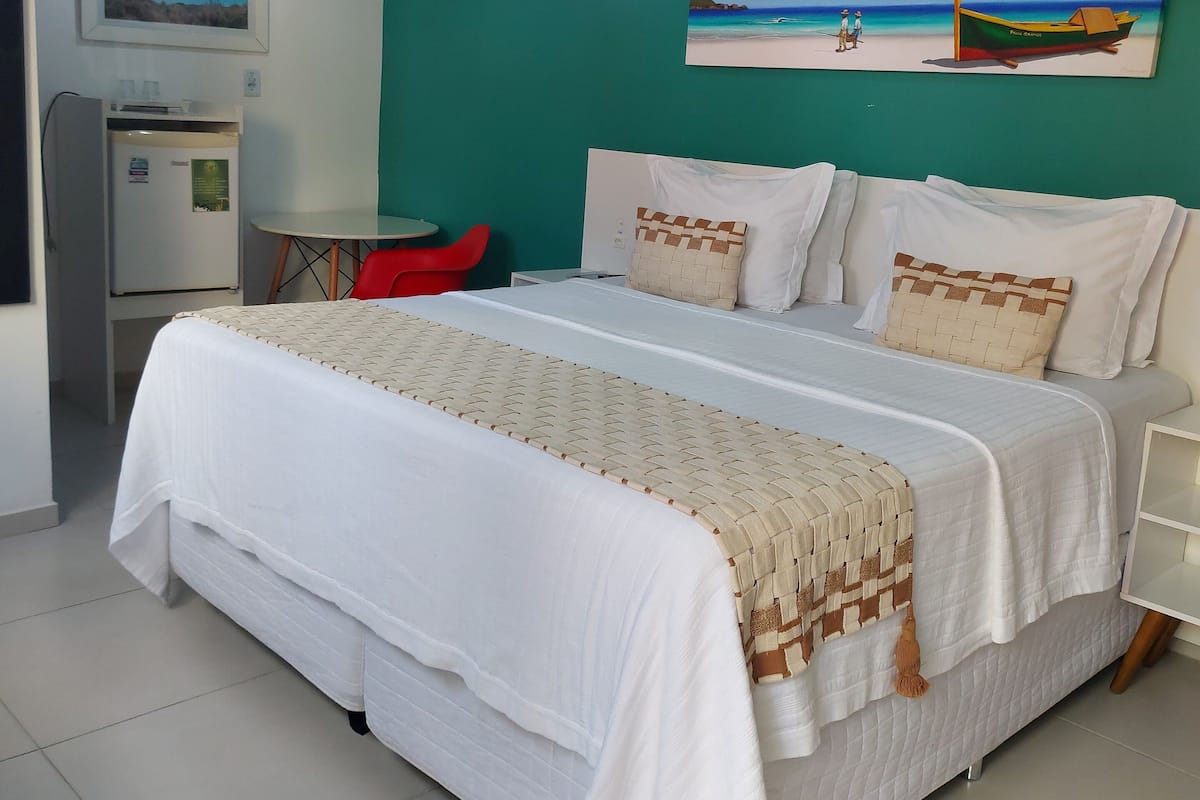 A neatly made bed with white bedding and a woven throw blanket, a small table with a red chair, a refrigerator, and a wall-mounted painting of a beach scene.