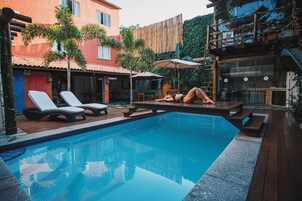 A swimming pool with lounge chairs and umbrellas, surrounded by buildings and greenery.