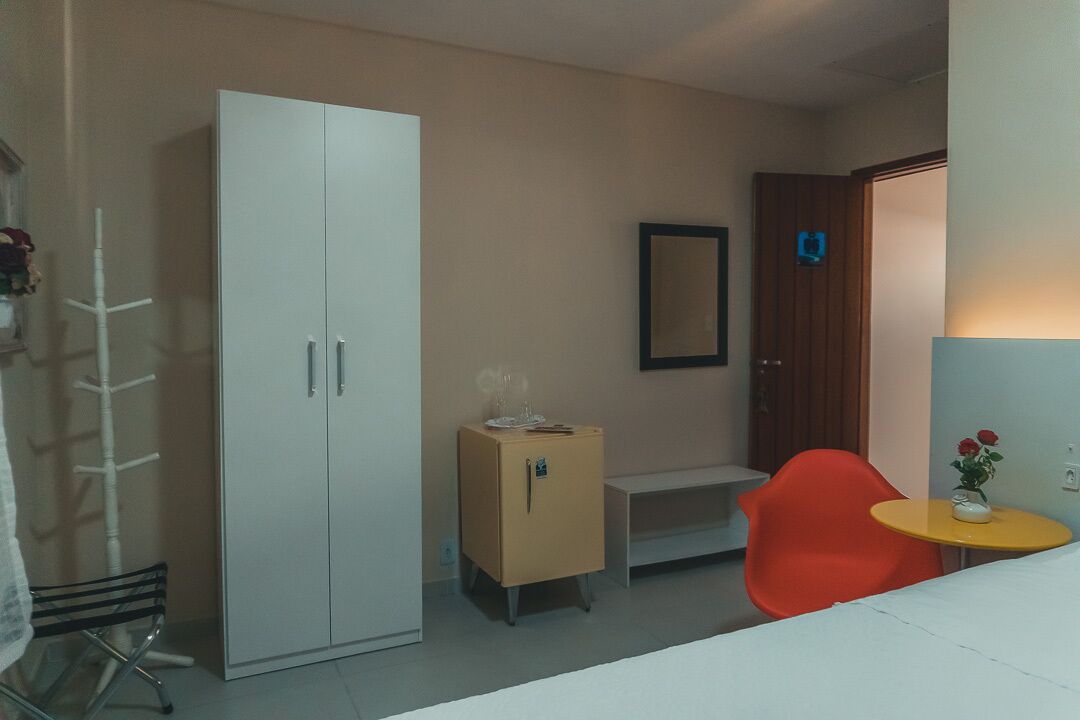 Una habitación con un armario blanco, un pequeño mueble, un espejo, una silla roja y una mesita con un jarrón.