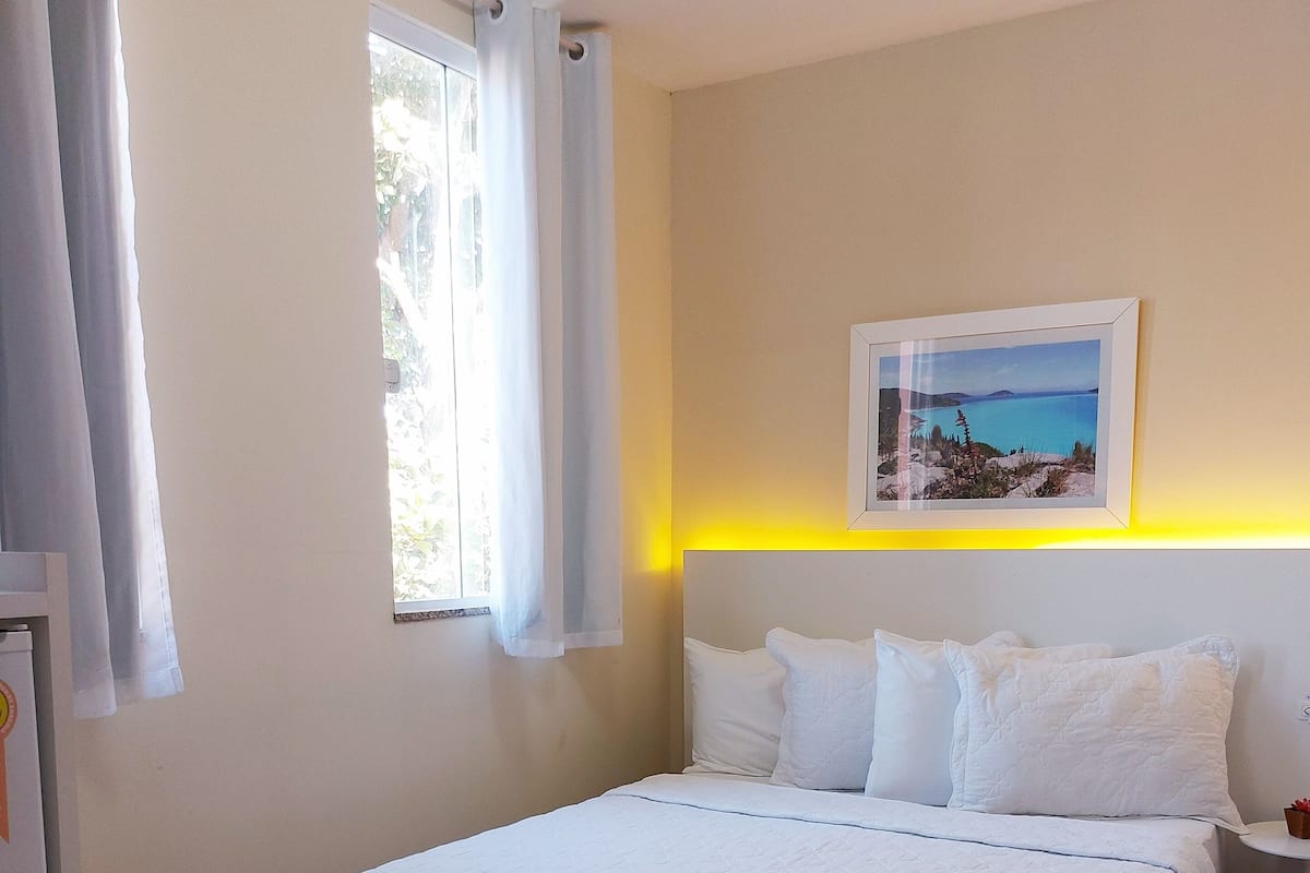 A bedroom with a bed, white curtains, a window, a nightstand, and a framed picture on the wall.