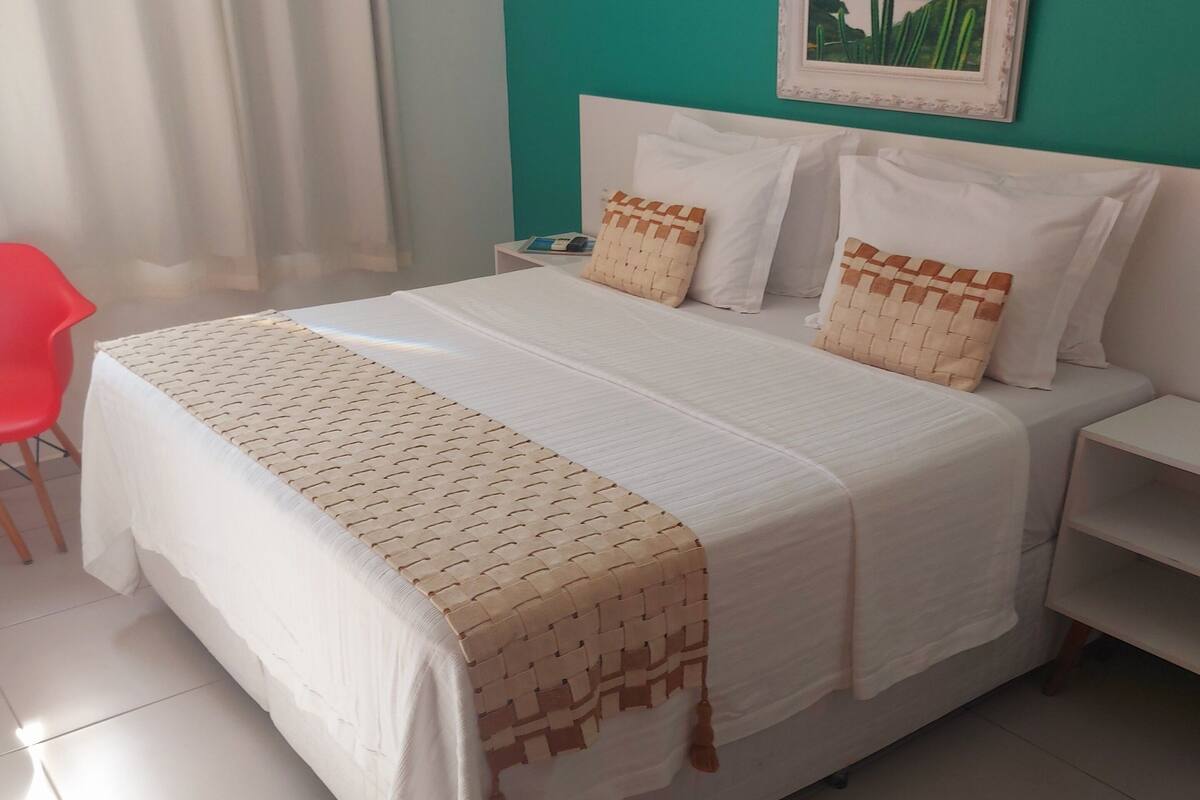 A neatly made bed with a patterned bedspread and pillows, a small bedside table with a book, and a painting on the wall.
