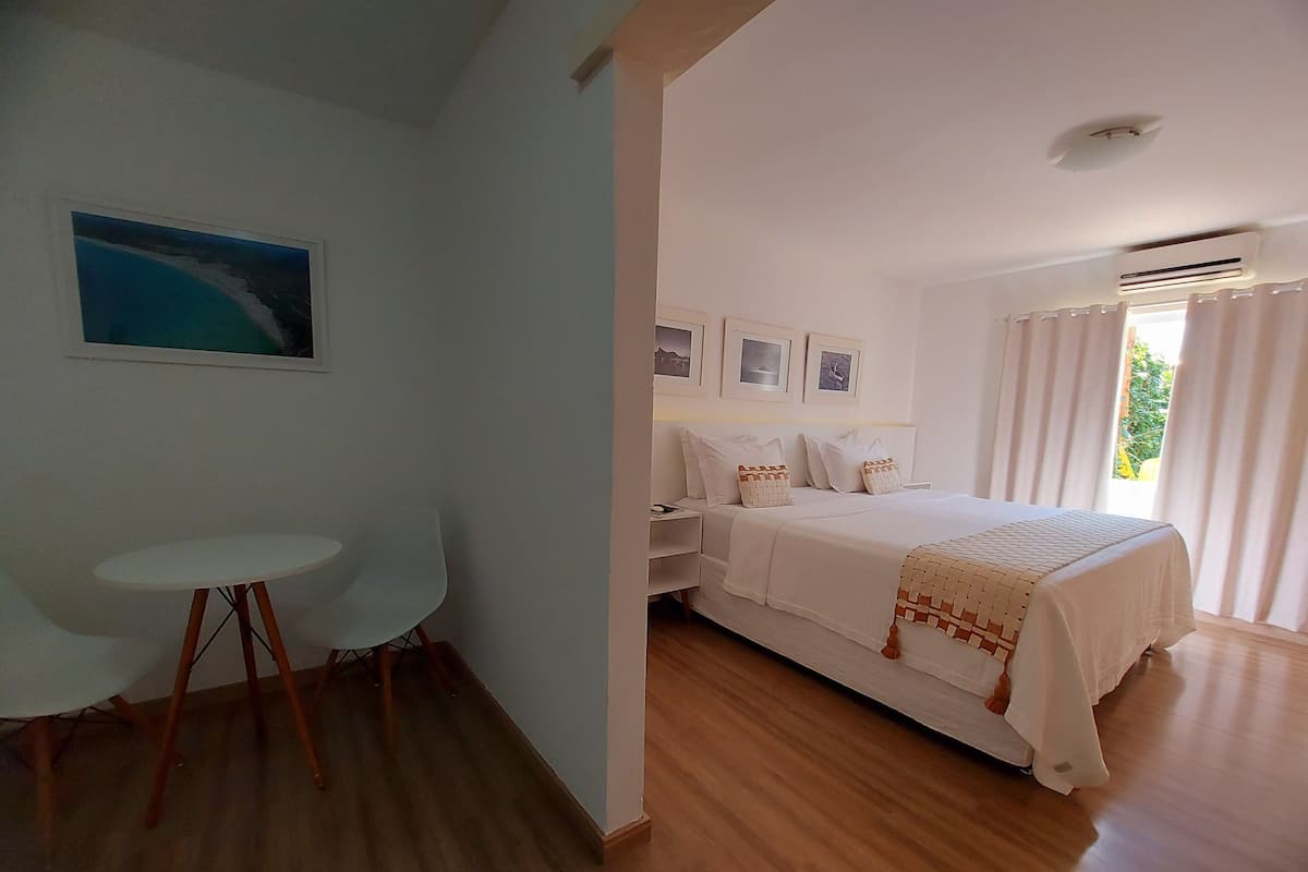 A bedroom with a bed, a small white table, and a chair. There is a window with curtains and a picture on the wall.