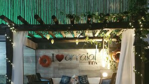 Un área de estar al aire libre con un banco de madera, un banco acolchado y una mesa pequeña. Se observa una pared con el texto "I love aronia inn" y elementos decorativos como un corazón y un pez.