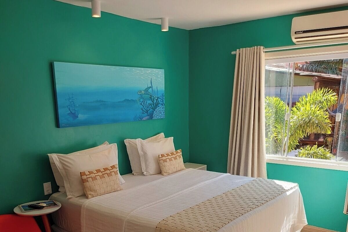 A bedroom with a bed, a small table, a red chair, a painting, and a window with curtains.