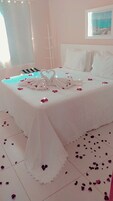 Una cama impecable con una toalla dispuesta en forma de corazón y pétalos de rosa esparcidos alrededor.