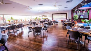 A spacious restaurant with multiple dining areas, ceiling fans, and a large screen displaying a presentation. - Hotel Menino Executive (Ponda)