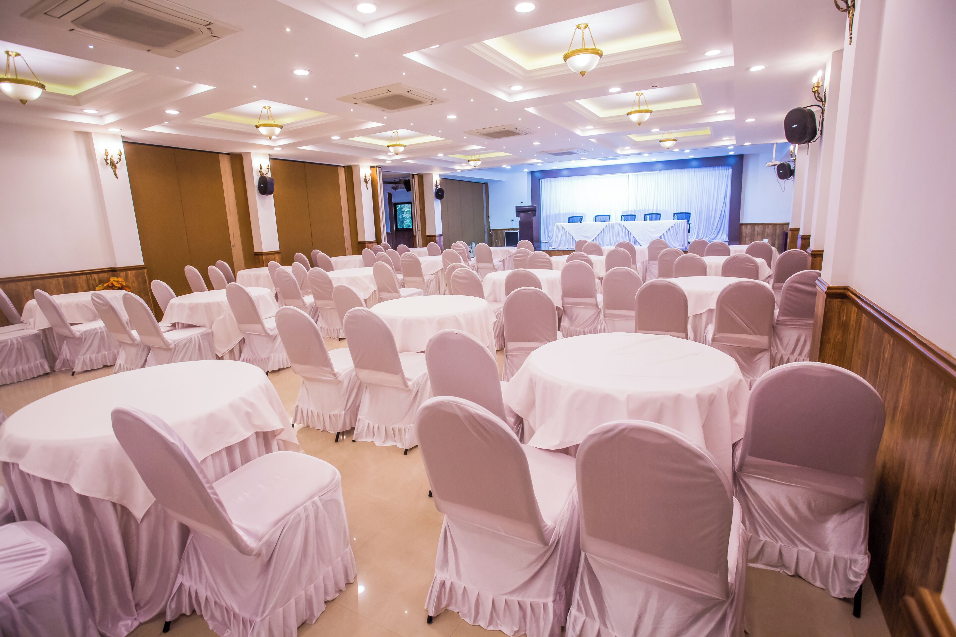 Uma sala de conferências com várias mesas redondas e cadeiras, equipada com luminárias no teto e alto-falantes.