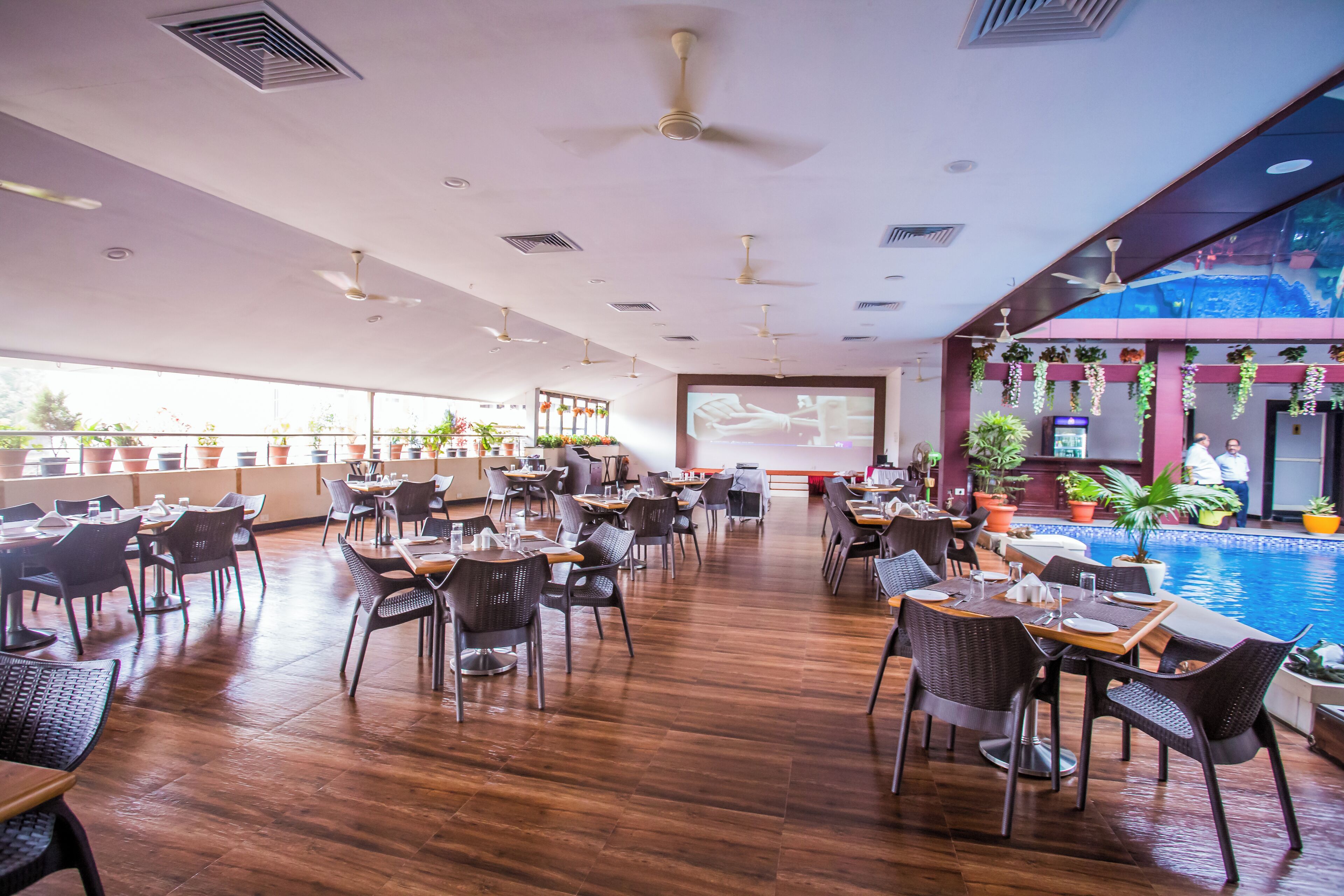 A spacious indoor area with multiple dining tables and chairs, a large screen displaying a video, and a pool area with potted plants.