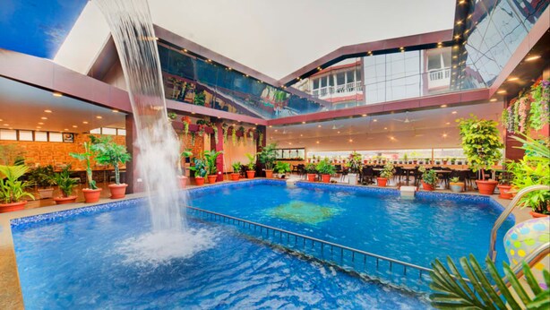 A large indoor pool with a waterfall feature, surrounded by potted plants and multiple levels of balconies. - Hotel Menino Executive (Ponda)