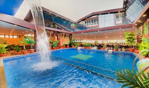 A large indoor pool with a waterfall feature, surrounded by potted plants and multiple levels of balconies. - Hotel Menino Executive (Ponda)