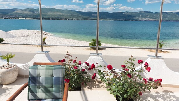 Une terrasse avec vue sur un lac et des montagnes, une chaise avec un coussin à carreaux et une plante en pot aux fleurs rouges.