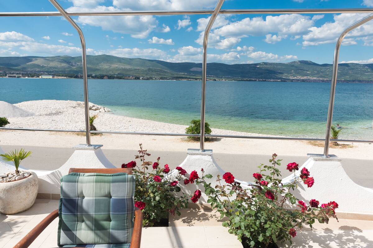 A terrace with a view of a lake and mountains, a chair with a plaid cushion, and a potted plant with red flowers.