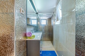 Une salle de bain moderne dotée d’une douche à parois de verre, d’un lavabo avec un miroir et d’une fenêtre laissant entrer la lumière naturelle.