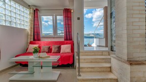 Une chambre moderne avec un canapé rouge, une table basse en verre et une vue sur la mer grâce à de grandes fenêtres.