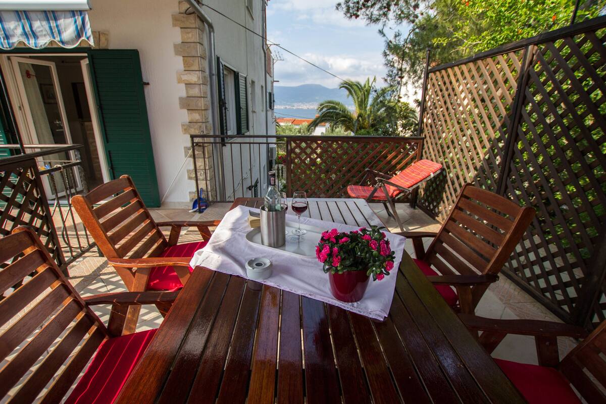 An outdoor wooden table with chairs, a potted plant, a bottle, and a glass of red wine on a white tablecloth.