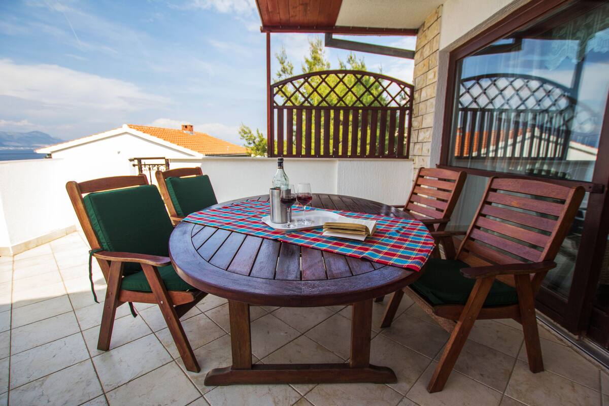 A round wooden table with four chairs, a metal bottle, and two wine glasses on a checkered tablecloth.