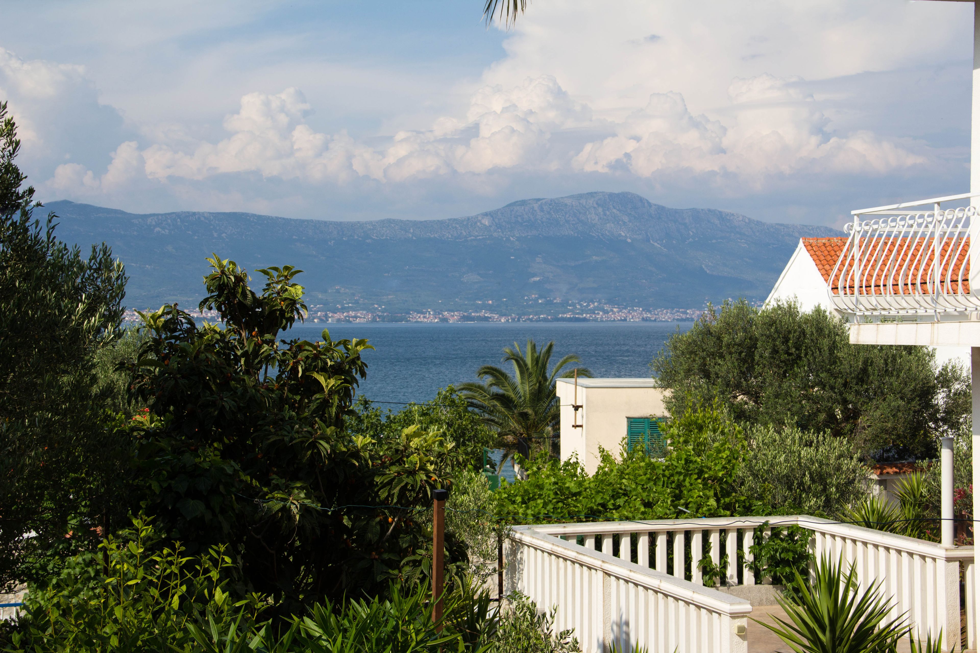 A coastal view with mountains in the background, lush greenery, and a white building with a red-tiled roof.