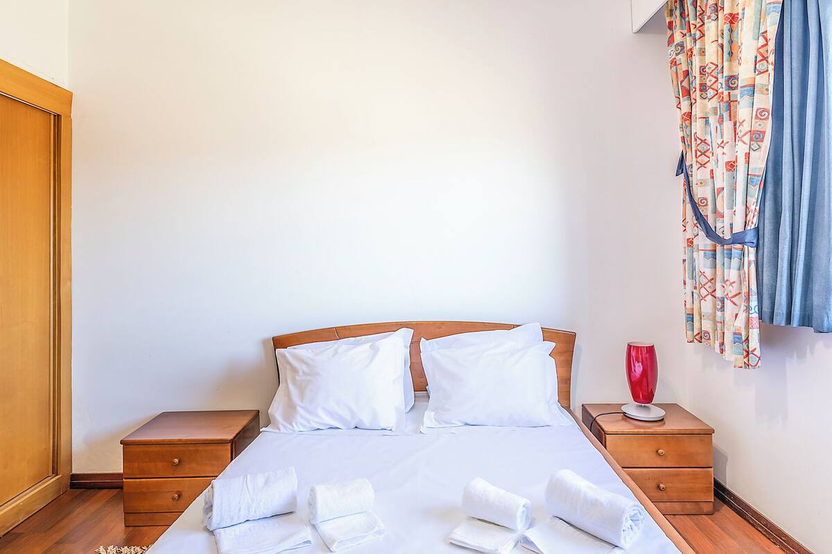 A neatly made bed with white linens, two wooden nightstands, a red vase, and patterned curtains.