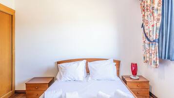 A neatly made bed with white linens, two wooden nightstands, a red vase, and patterned curtains.